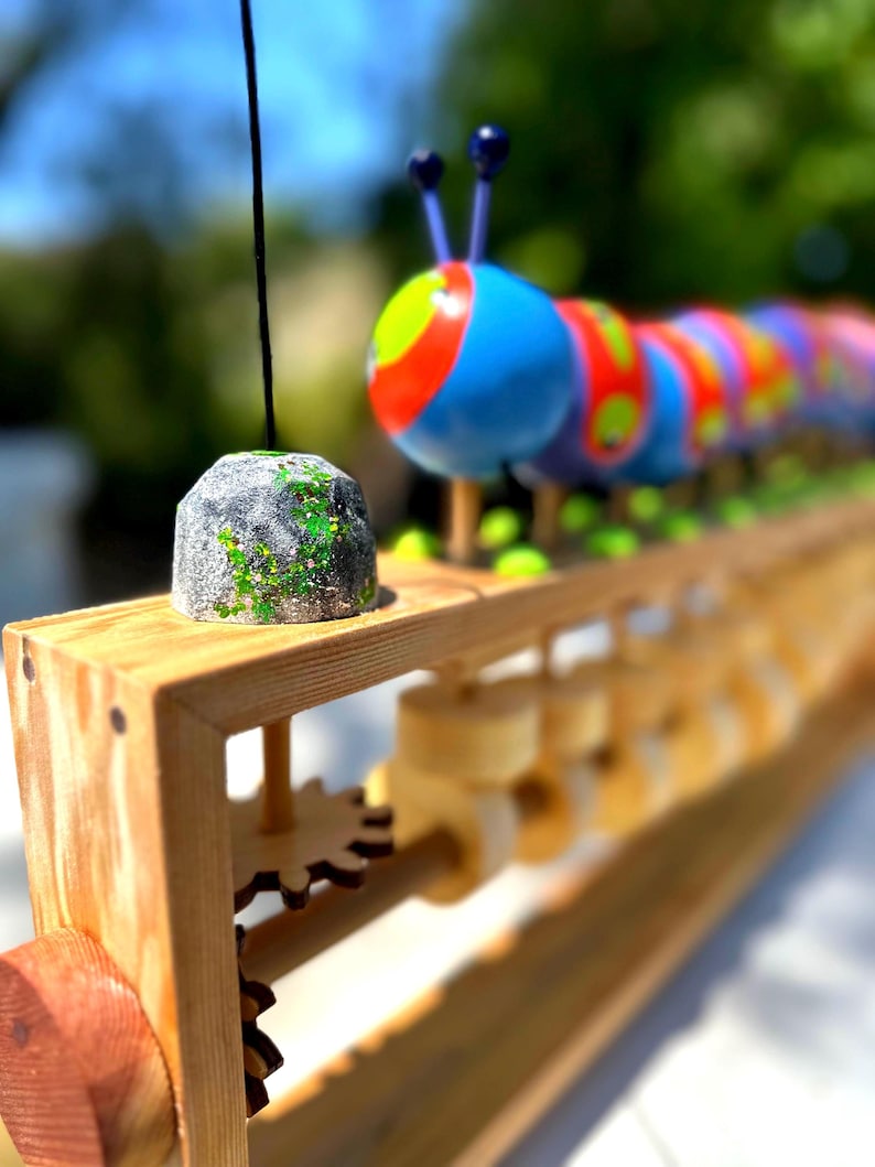 May include: A wooden toy featuring a colorful caterpillar with blue, red, yellow, and green segments. The caterpillar sits on a wooden track with gears and a small, gray, rock-like piece. The toy is designed with a focus on bright colors and playful design.