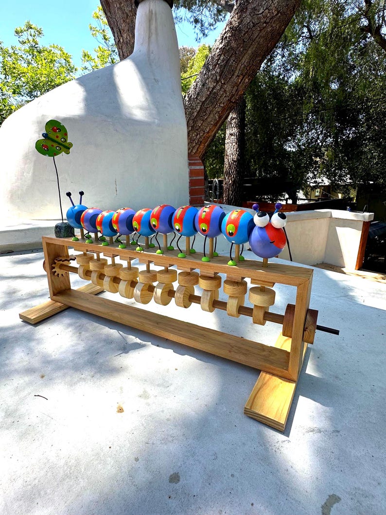 May include: A whimsical wooden caterpillar sculpture with colorful, round segments. Each segment is painted in vibrant hues of blue, red, and yellow, with large, googly eyes. The sculpture sits on a wooden frame with turning gears, creating a playful, kinetic art piece.