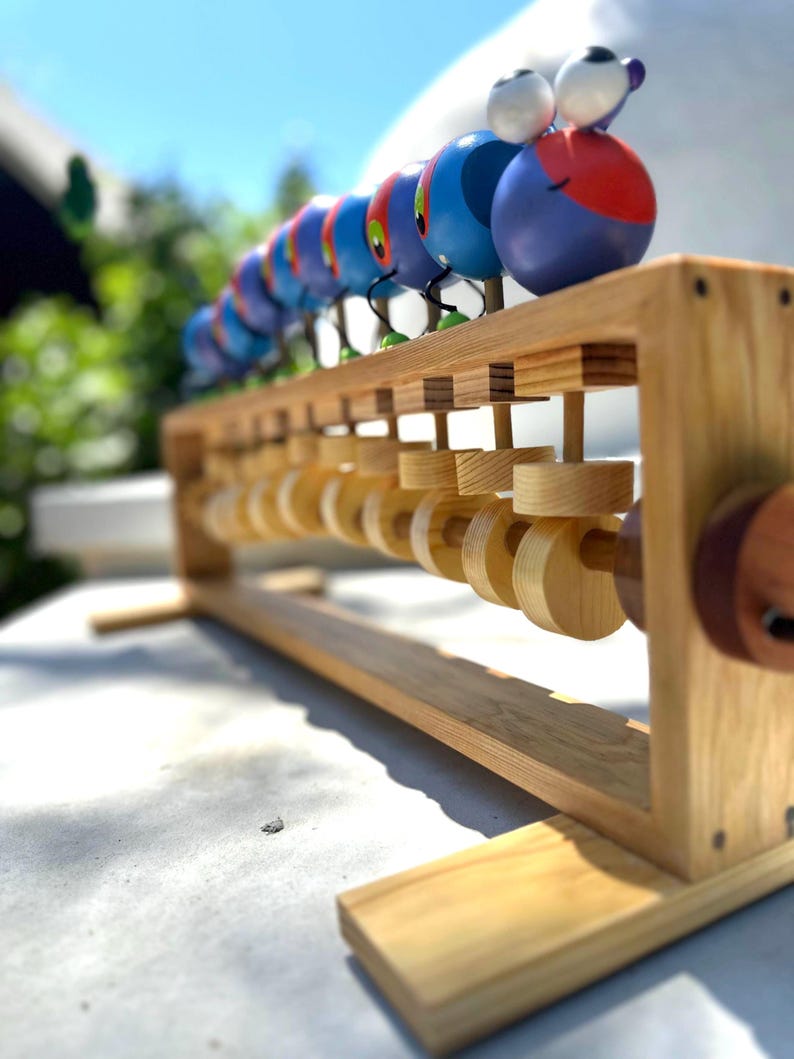 May include: A wooden abacus with a colorful caterpillar design. The caterpillar is made of painted wooden spheres in shades of blue, red, and purple. The abacus frame is made of light-colored wood, with wooden beads for counting.