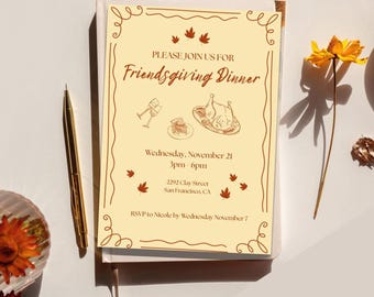 May include: A Friendsgiving Dinner invitation on a stack of books. The invitation is cream-colored with brown text and illustrations of food and fall leaves. A gold pen and a yellow flower are next to the invitation.