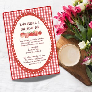 Op de afbeelding: Uitnodiging voor een bessenplukdag met een rood-wit geruit patroon. De ovale kaart toont aardbeien en tekst. Een boeket roze en witte bloemen en een glas koffie staan op de achtergrond.