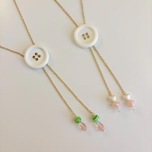 May include: Two gold-tone chain necklaces featuring a central white four-hole button. One necklace ends with green striped beads and pink teardrop charms, while the other features white heart beads and faceted pink spherical beads at the ends.