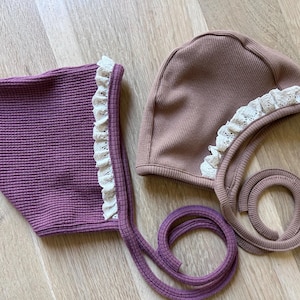 May include: Two baby bonnets. One is a deep purple with a waffle texture and white lace trim. The other is a muted rose color with a ribbed texture and white lace trim. Both bonnets have ties and are made of a soft material.