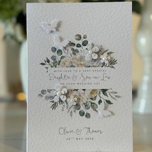 Może przedstawiać: Biała kartka ślubna z kwiatowymi i motylkowymi zdobieniami. Na kartce widnieje napis "WITH LOVE TO A VERY SPECIAL Daughter & Son-in-Law ON YOUR WEDDING DAY" oraz imiona "Olivia & Thomas" i data "24 maja 2026".