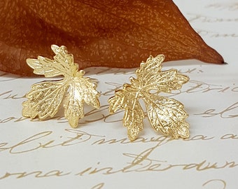 Puede incluir: Un par de pendientes de hoja de oro. Los pendientes están hechos de metal texturizado y tienen una forma delicada y orgánica.