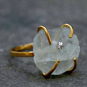 May include: A gold-toned ring featuring a rough-cut, pale blue gemstone with a small, sparkling accent stone. The ring's band and prongs are a bright gold color, creating a striking contrast with the stone.