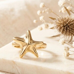 Könnte beinhalten: Ein goldfarbener Seesternring. Der Ring liegt auf einer weißen Marmoroberfläche, im Hintergrund getrocknete Blumen. Das Seestern-Design ist der Mittelpunkt, mit fünf ausgeprägten Spitzen. Der Ring ist ein auffälliges Schmuckstück.