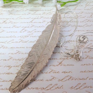 May include: A silver feather necklace with a silver charm pendant. The feather is detailed and textured, and the charm is a small, stylized letter.