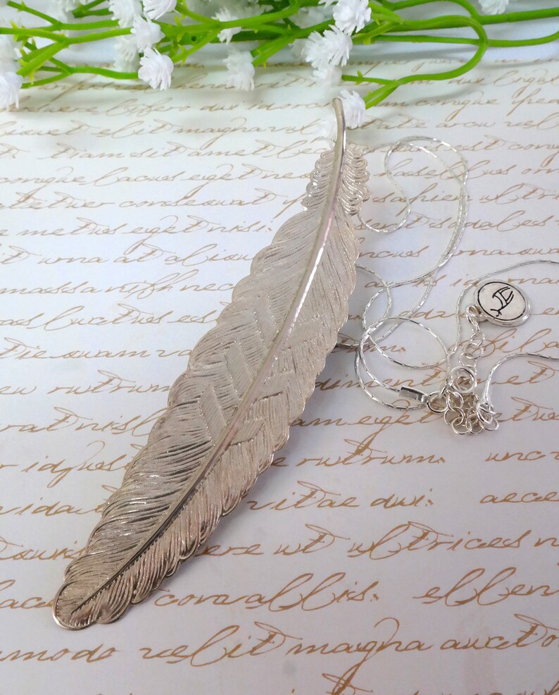 May include: A silver feather necklace with a silver charm pendant. The feather is detailed and textured, and the charm is a small, stylized letter.