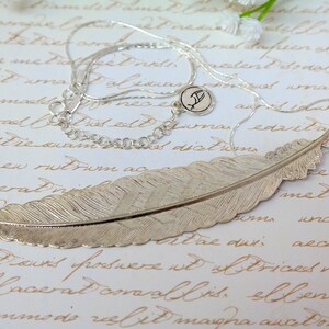 May include: A silver feather pendant necklace with a delicate chain and a small charm with the letter 'F'. The feather pendant is detailed and realistic, with a textured surface.