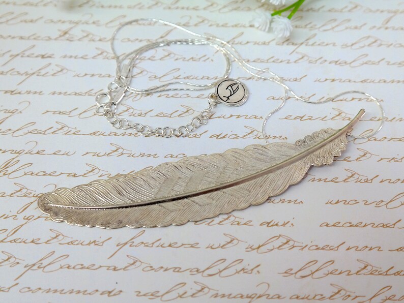 May include: A silver feather pendant necklace with a delicate chain and a small charm with the letter 'F'. The feather pendant is detailed and realistic, with a textured surface.
