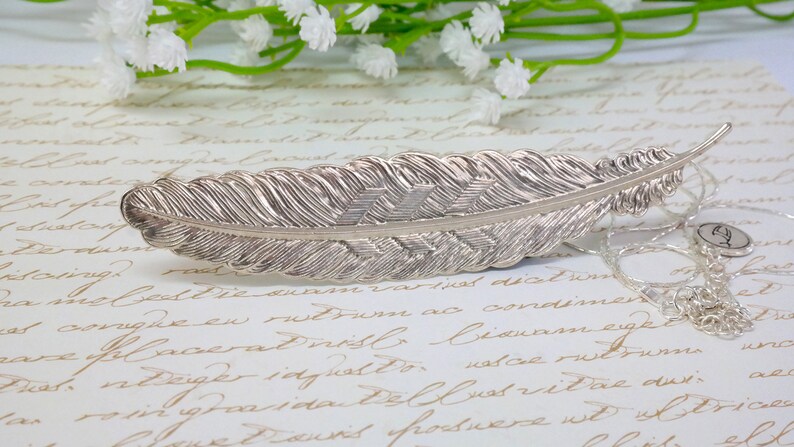 May include: A silver feather pendant necklace with a delicate chain and a small charm. The feather is detailed with a textured design.