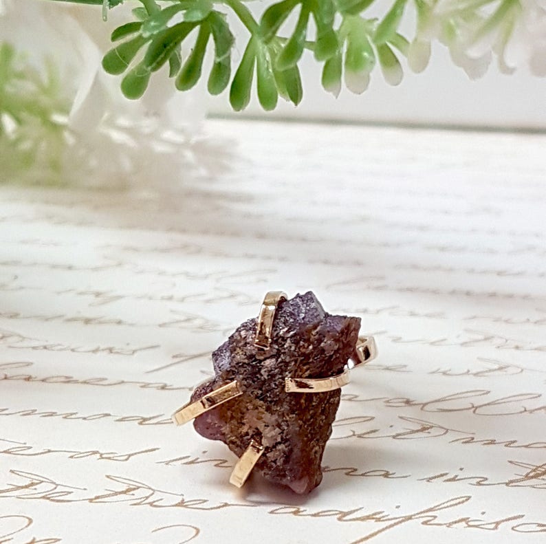 May include: A ring featuring a raw, purple amethyst stone set in a gold band. The stone is irregularly shaped and has a rough, natural texture.