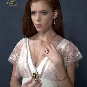 Puede incluir: Una mujer con un vestido blanco de encaje con un collar y pendientes de hoja de oro. El collar tiene un peque&ntilde;o dije de hoja de oro. La mujer lleva un anillo de oro en su mano derecha. El fondo es azul oscuro.
