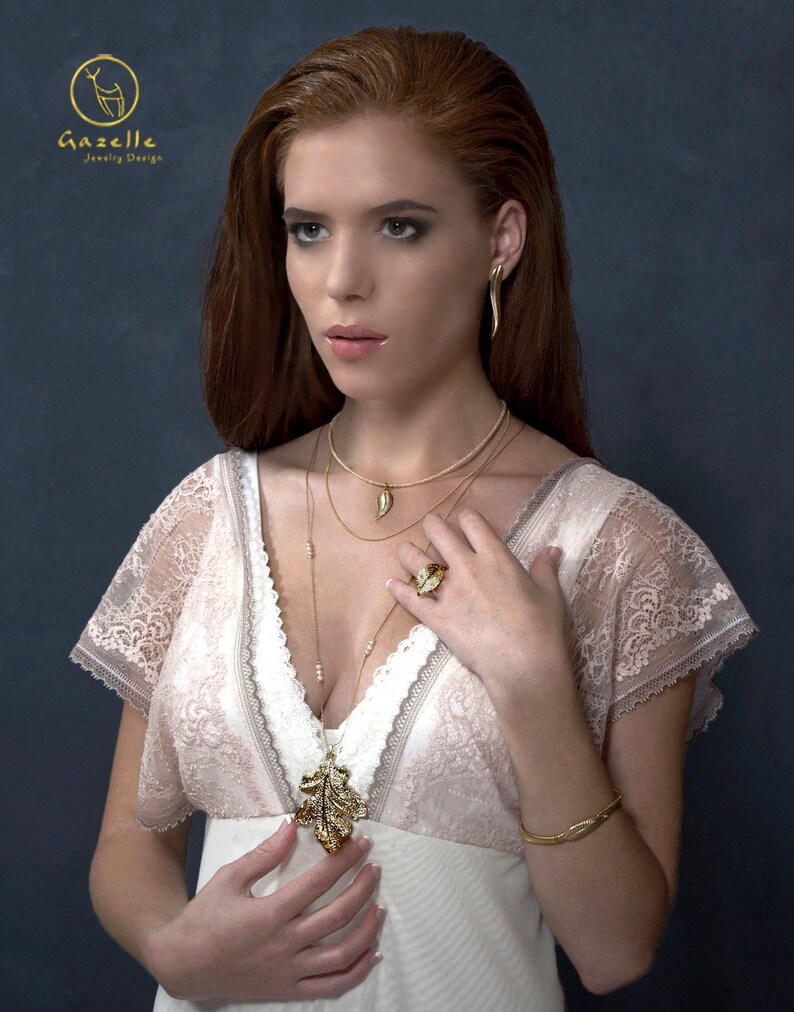 Puede incluir: Una mujer con un vestido blanco de encaje con un collar y pendientes de hoja de oro. El collar tiene un peque&ntilde;o dije de hoja de oro. La mujer lleva un anillo de oro en su mano derecha. El fondo es azul oscuro.