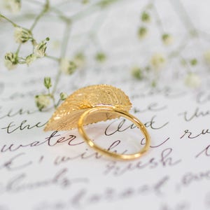 Puede incluir: Un anillo de hoja de oro con un dise&ntilde;o delicado. El anillo est&aacute; apoyado en un trozo de papel con texto escrito a mano.
