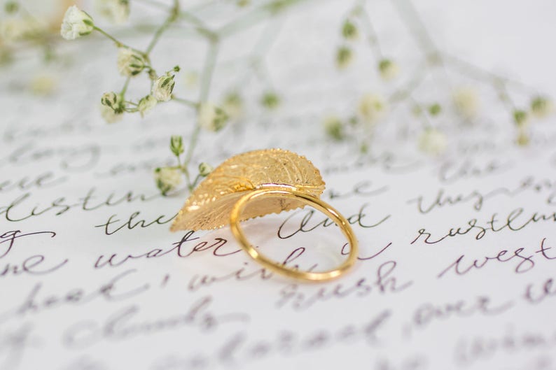 Puede incluir: Un anillo de hoja de oro con un dise&ntilde;o delicado. El anillo est&aacute; apoyado en un trozo de papel con texto escrito a mano.