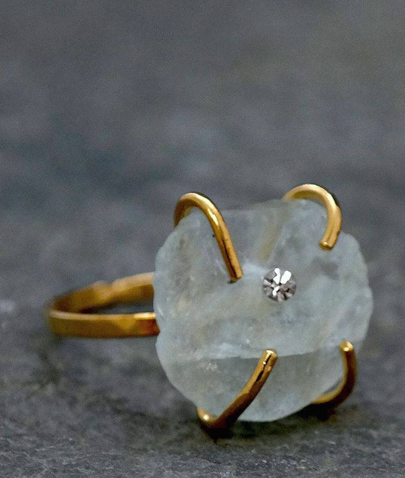 May include: A gold-toned ring featuring a rough-cut, pale blue gemstone with a small, sparkling accent stone. The ring's band and prongs are a bright gold color, creating a striking contrast with the stone.