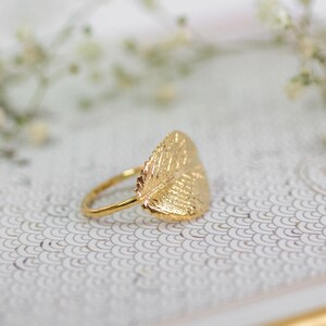 Puede incluir: Un anillo de oro con un dise&ntilde;o de hoja. El anillo est&aacute; sobre una superficie blanca con un patr&oacute;n.