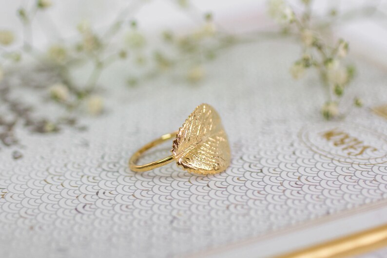 Puede incluir: Un anillo de oro con un dise&ntilde;o de hoja. El anillo est&aacute; sobre una superficie blanca con un patr&oacute;n.