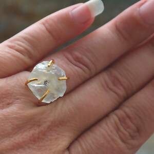 May include: A gold-toned ring featuring a rough-cut, pale-yellow gemstone with a small, clear stone at its center. The ring's setting has four gold prongs that secure the stone. The ring is shown on a hand.