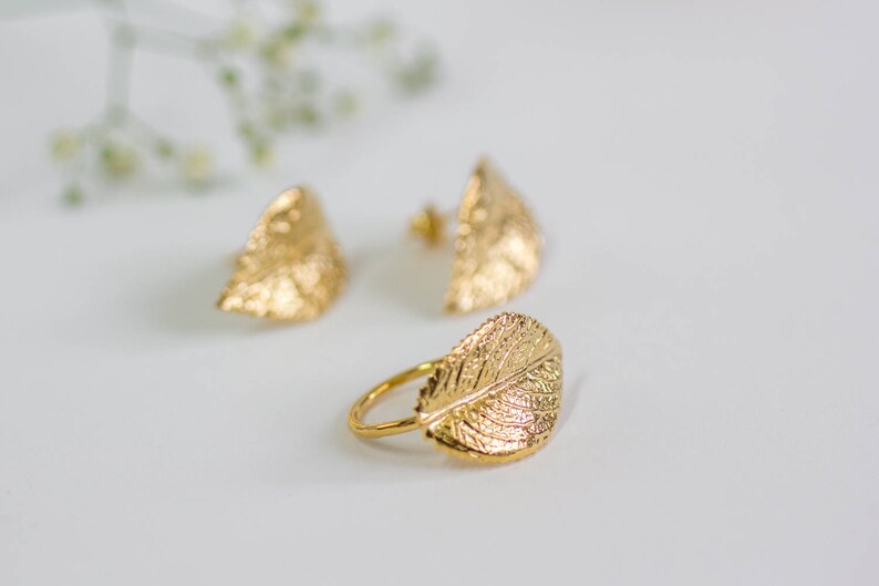 Puede incluir: Un conjunto de pendientes y un anillo de hoja de oro. Los pendientes tienen forma de hoja y una superficie texturizada. El anillo tambi&eacute;n tiene forma de hoja y una superficie texturizada.