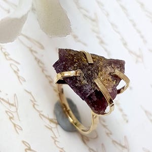 May include: A gold ring with a large, raw, purple and gold gemstone set in a simple, modern design.