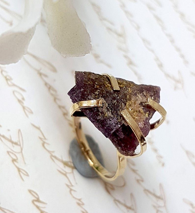 May include: A gold ring with a large, raw, purple and gold gemstone set in a simple, modern design.