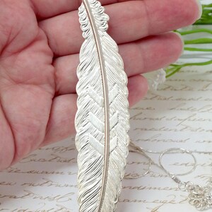 May include: A silver feather pendant necklace with a detailed design. The feather is textured and has a subtle shine. The necklace is on a delicate chain.