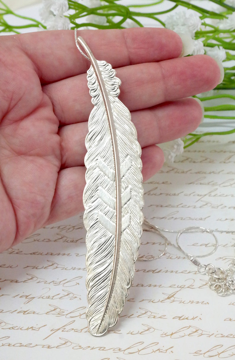 May include: A silver feather pendant necklace with a detailed design. The feather is textured and has a subtle shine. The necklace is on a delicate chain.