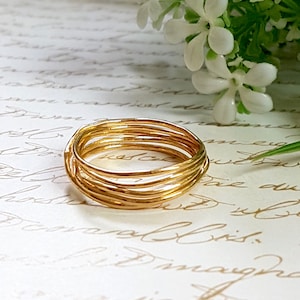 Puede incluir: Un anillo dorado hecho de múltiples bandas delgadas retorcidas juntas. El anillo está sobre una superficie blanca con flores blancas en el fondo.
