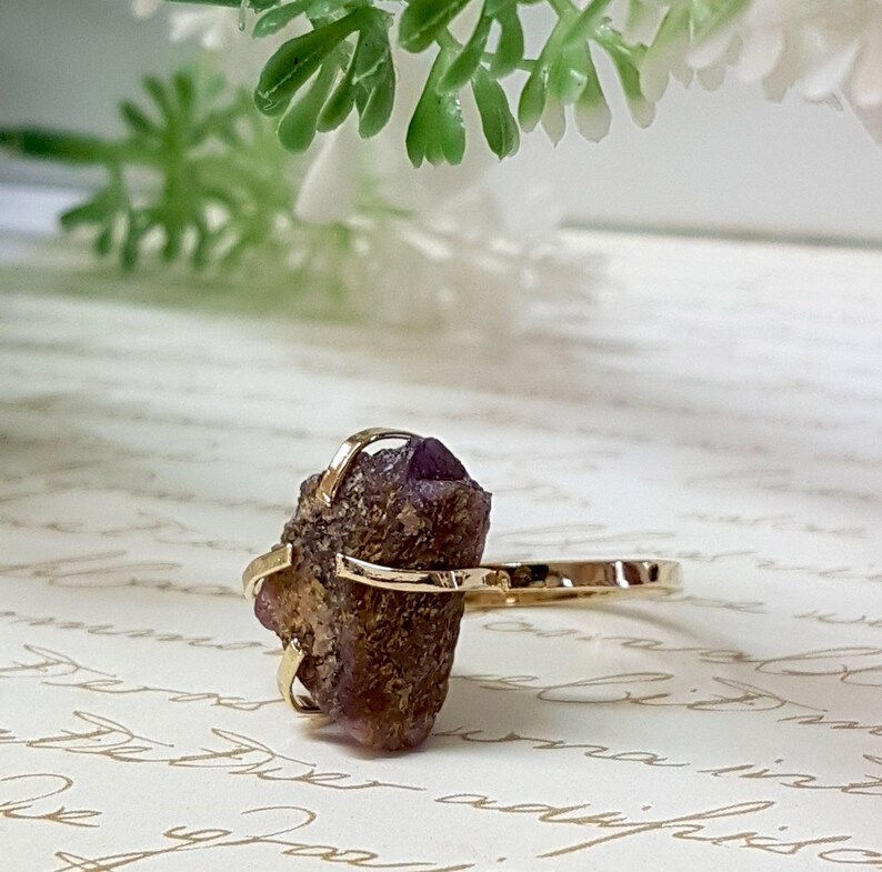 May include: A gold ring with a raw amethyst stone set in a delicate wire cage. The stone is a deep purple color and has a rough, natural texture.