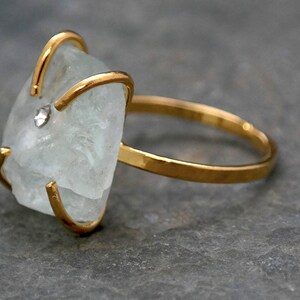 May include: A gold-toned ring featuring a large, irregular-shaped, pale blue gemstone. The gemstone is secured by gold prongs and a small, clear accent stone. The ring band is a simple, polished gold. The ring is displayed on a gray surface.