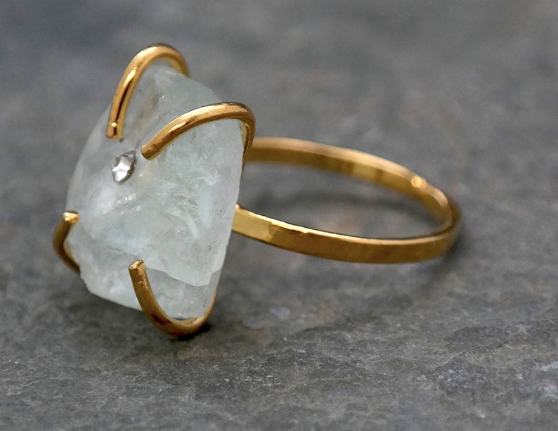 May include: A gold-toned ring featuring a large, irregular-shaped, pale blue gemstone. The gemstone is secured by gold prongs and a small, clear accent stone. The ring band is a simple, polished gold. The ring is displayed on a gray surface.