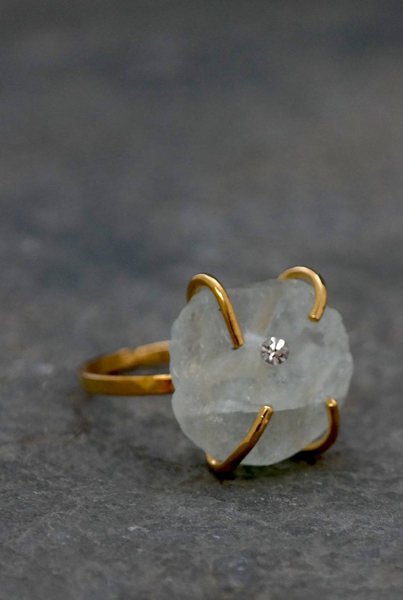 May include: A gold-toned ring featuring a rough-cut, pale blue gemstone with a small, sparkling accent stone. The gemstone is held in place by four gold prongs. The ring's band is also gold, creating a cohesive and elegant design.