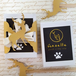 May include: Two gift boxes with a gold deer design. The box on the left has a white ribbon and a gold paw print. The box on the right has a black background with a gold deer silhouette and the text "Gazelle" in gold. The text "כלב, חתול, וציפור" is in Hebrew below the word "Gazelle". A white paw print is below the text.