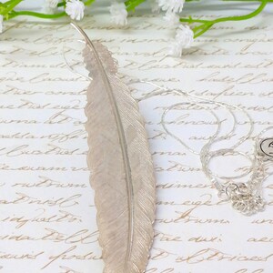 May include: A silver feather pendant necklace with a delicate chain. The feather is detailed and has a textured surface. The necklace is displayed on a white background with a handwritten note.