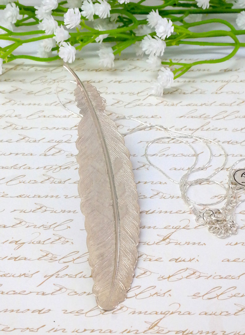 May include: A silver feather pendant necklace with a delicate chain. The feather is detailed and has a textured surface. The necklace is displayed on a white background with a handwritten note.