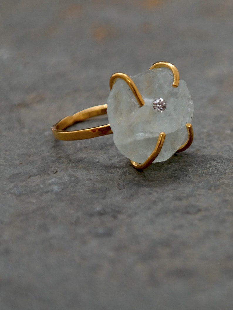 May include: A gold ring featuring a light blue, irregularly shaped gemstone with a small, round, clear stone in the center. The gemstone is held in place by four gold prongs. The ring band is also gold.
