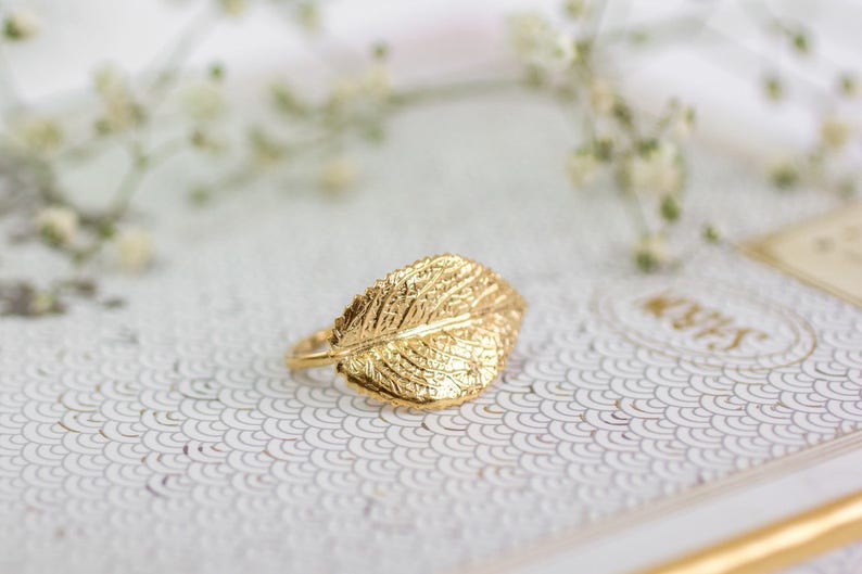 Puede incluir: Un anillo de hoja de oro con un dise&ntilde;o detallado. El anillo est&aacute; sobre una superficie blanca con un patr&oacute;n.