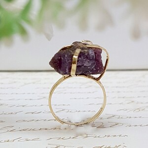 May include: A gold ring with a large, raw, purple gemstone set in the center. The gemstone is held in place by a gold wire frame.