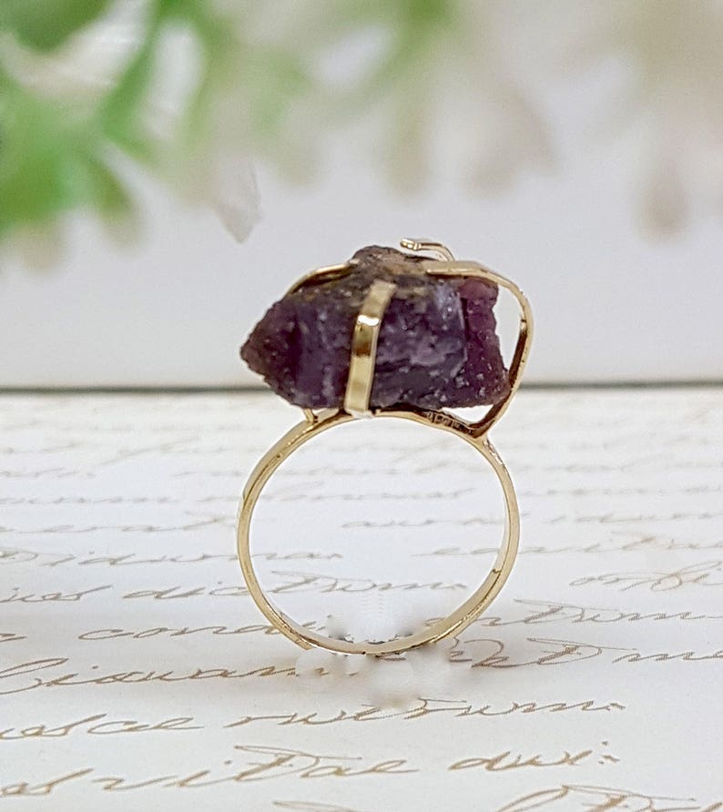 May include: A gold ring with a large, raw, purple gemstone set in the center. The gemstone is held in place by a gold wire frame.