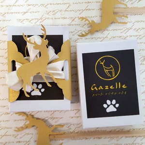 May include: Two small gift boxes with gold accents. The box on the left has a gold deer cutout with a white ribbon. The box on the right has a black background with a gold deer logo and the text "Gazelle" in gold. The text "כלב (כלב) ורוד" is in Hebrew below the logo.