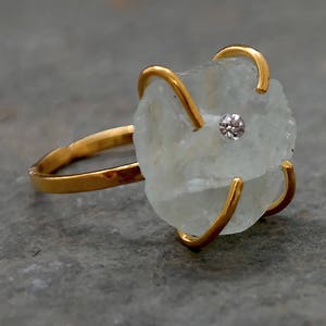 May include: A gold ring featuring a rough-cut, pale blue gemstone with a small, clear stone at its center. The gold band and prongs cradle the irregular stone, creating a unique and organic design. The ring is set against a gray background.