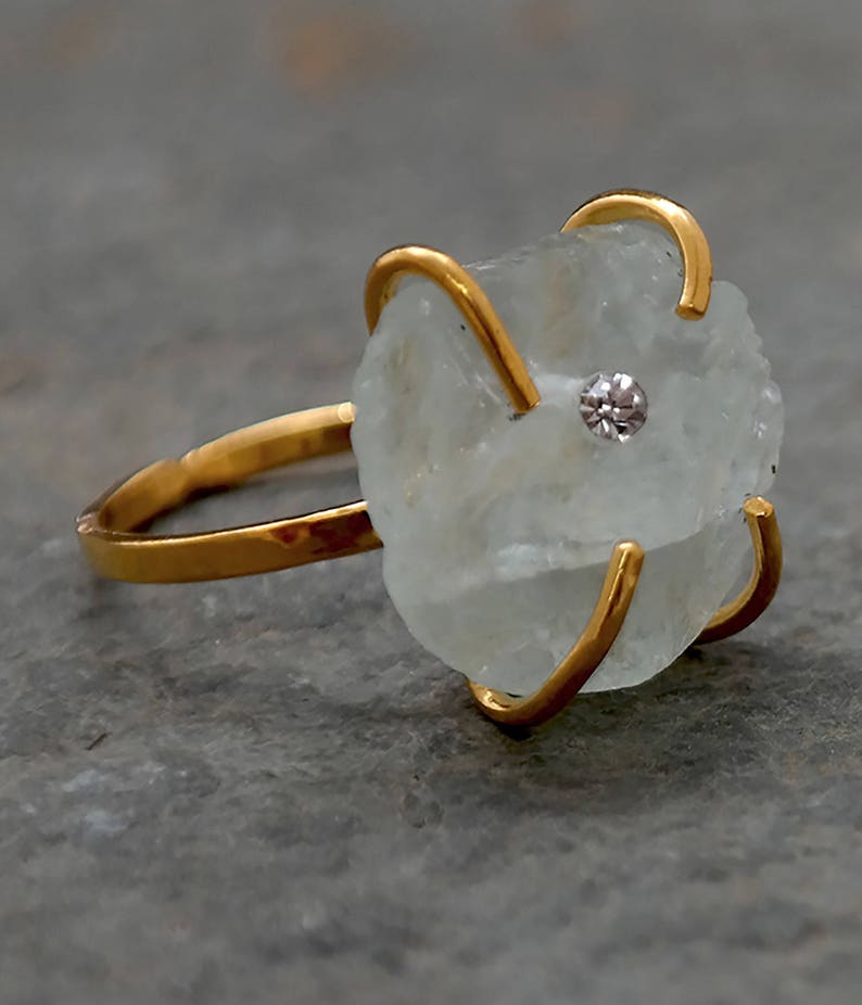 May include: A gold ring featuring a rough-cut, pale blue gemstone with a small, clear stone at its center. The gold band and prongs cradle the irregular stone, creating a unique and organic design. The ring is set against a gray background.