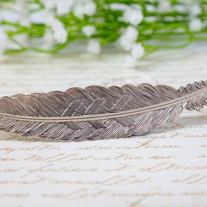 May include: A silver feather bookmark with a detailed etched design. The feather is curved and has a pointed tip.