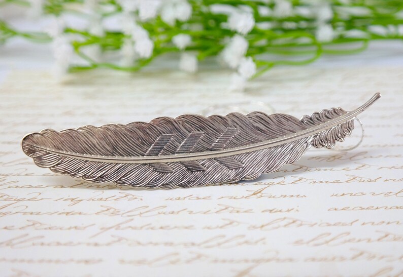 May include: A silver feather bookmark with a detailed etched design. The feather is curved and has a pointed tip.