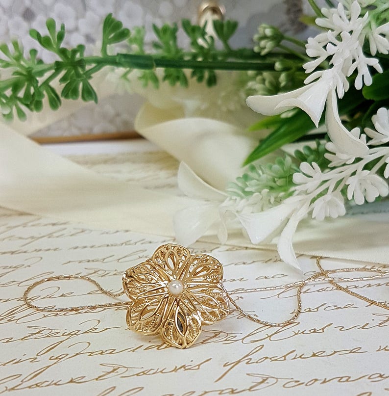 May include: A gold filigree flower pendant necklace with a small pearl in the center. The necklace is on a white surface with a floral background.