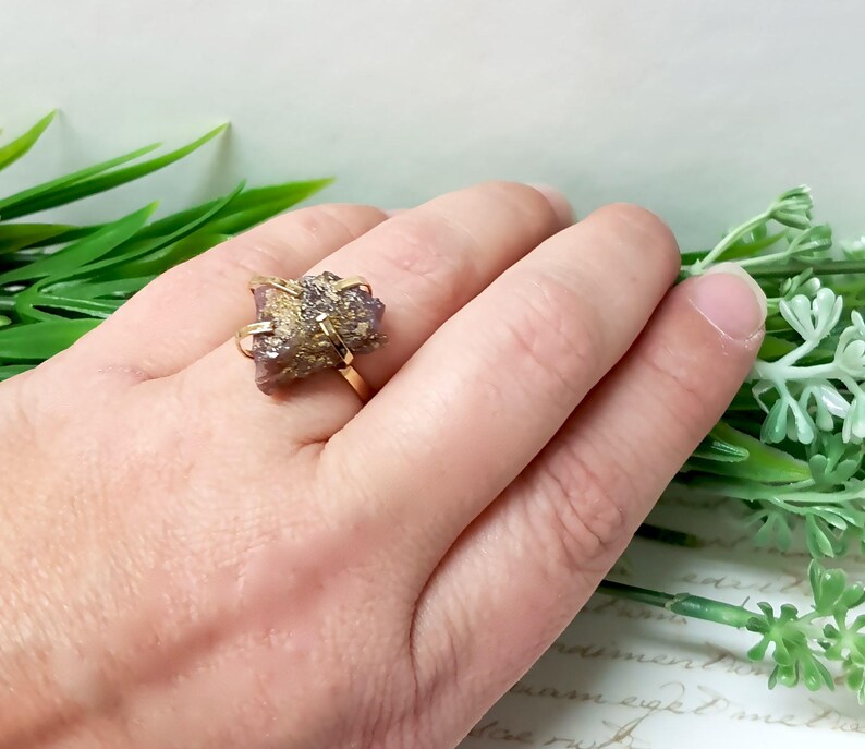 May include: A gold ring with a pink and gold crystal stone. The ring is on a hand.