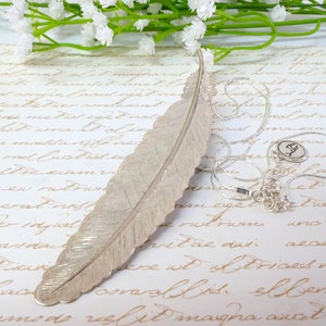 May include: A silver feather bookmark with a delicate, textured surface. The bookmark is lying on a piece of paper with handwritten text.